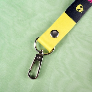 May include: A lanyard with a yellow and black strap. The black portion features a repeating pattern of yellow skulls and pink floral designs. The lanyard has a silver-colored metal clasp and a snap closure.