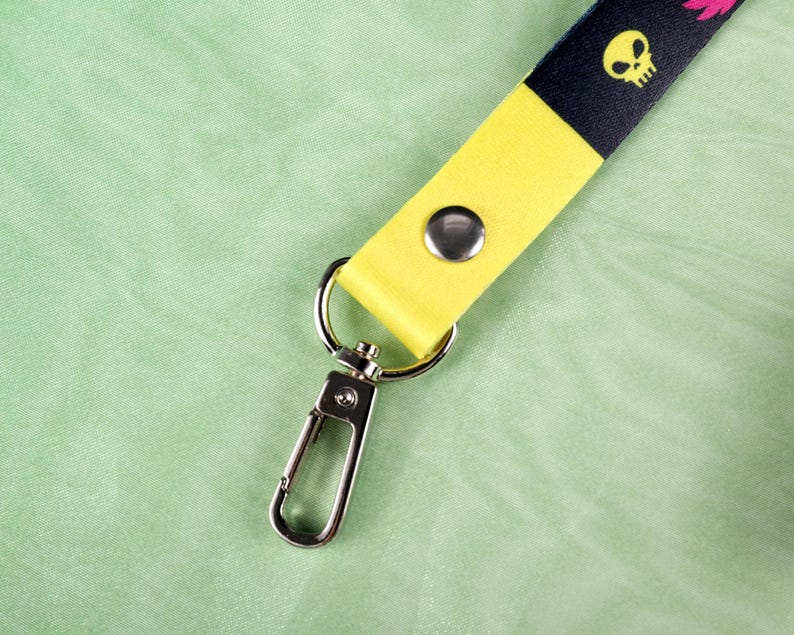 May include: A lanyard with a yellow and black strap. The black portion features a repeating pattern of yellow skulls and pink floral designs. The lanyard has a silver-colored metal clasp and a snap closure.