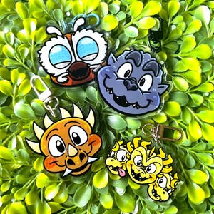 May include: Four colorful cartoon character keychains with clear plastic backing. The characters are a yellow and orange dinosaur, a purple monster, a yellow and orange three-headed dragon, and a white and orange bee.