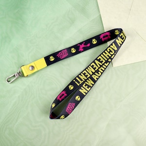 May include: Black lanyard with yellow accents and silver clasp. The lanyard features pink and yellow skull and crown graphics, and the words "NEW ACHIEVEMENT!" in yellow.