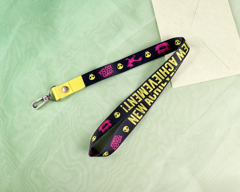 May include: Black lanyard with yellow accents and silver clasp. The lanyard features pink and yellow skull and crown graphics, and the words "NEW ACHIEVEMENT!" in yellow.