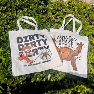 May include: Two white canvas tote bags with handles. The left bag features a cartoon character running with the text DIRTY DIRTY DIRTY and WHY ROOM SO MESSY, QUESTION? The right bag shows a brown robot character with the text AMAZE AMAZE AMAZE.
