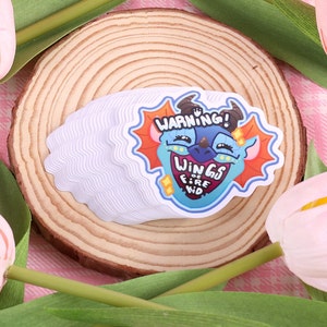 May include: A stack of white stickers with a cartoon dragon face on top. The dragon is blue with orange wings and a red mouth. The sticker says "Warning! Wings of Fire Kid".