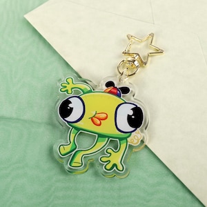 May include: A novelty frog keyring with a gold star-shaped clasp. The frog is yellow and green with large black eyes, a red and orange mouth, and a small hat. The keyring is on a light green surface with a white envelope.
