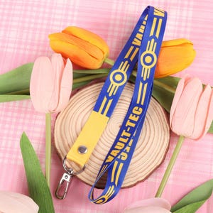 May include: A blue and yellow lanyard featuring the text "VAULT-TEC" and a circular logo. The lanyard is displayed on a wooden slice, with pink and yellow tulips surrounding it.