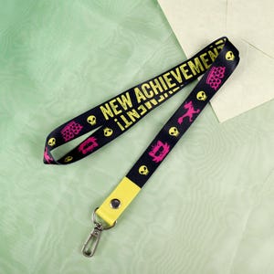 Może przedstawiać: Czarna smycz z żółtym napisem "NEW ACHIEVEMENT!" i powtarzającymi się różowymi i żółtymi grafikami czaszek, koron i innych kształtów. Smycz ma żółtą końcówkę ze srebrnym zapięciem.