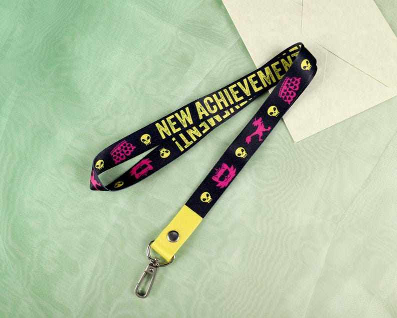 May include: Black lanyard with yellow text that reads "NEW ACHIEVEMENT!" and repeating pink and yellow graphics of skulls, crowns, and other shapes. The lanyard has a yellow end piece with a silver clasp.