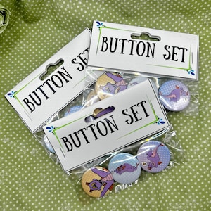 May include: Button sets featuring cartoon dragon designs. Each set includes several round buttons in clear packaging, with the words "BUTTON SET" printed on the top. The buttons have a variety of colors, including purple, yellow, and blue.