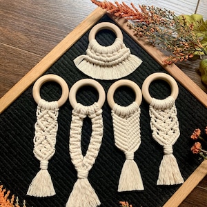May include: Five wooden ring teethers with macrame fringe and tassels. The teethers are displayed on a black background.