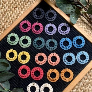 Könnte beinhalten: Eine Sammlung handgefertigter Makramee-Ohrringe in verschiedenen Farben, darunter Blau, Rosa, Gelb und Grün. Die Ohrringe werden auf einer schwarzen Oberfläche in einem Holzrahmen präsentiert, die ihr aufwendiges Webdesign zeigt.