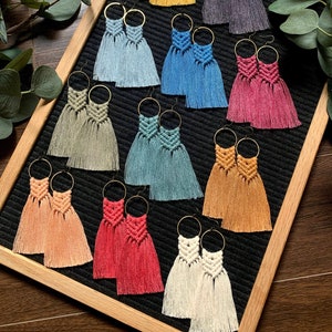 Puede incluir: Una colección de 10 pares de pendientes de macramé hechos a mano con aros dorados. Los pendientes se muestran sobre un fondo negro con un marco de madera. Los pendientes son de varios colores, incluyendo azul, rojo, verde, amarillo y blanco.