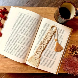 May include: A close-up of a beige macrame bookmark with a tassel. The bookmark is lying on an open book with a cup of coffee and a pine cone on a wooden surface.