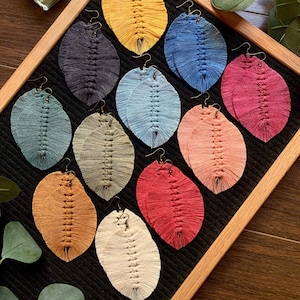 Puede incluir: Una muestra de diez pares de pendientes de macramé hechos a mano en varios colores, incluyendo amarillo, azul, verde, rojo y beige. Los pendientes se muestran sobre un fondo negro con un marco de madera.