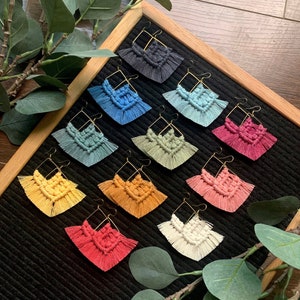 May include: A set of 10 pairs of handmade macrame earrings in various colors, including black, blue, green, yellow, pink, and white. The earrings are displayed on a black background with a wooden frame.