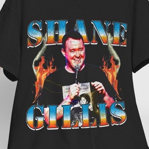 May include: Black t-shirt featuring a graphic of a man holding a microphone, with the name "SHANE GILLIS" in a vintage-style font. The design includes flames and a portrait of the comedian. A white hat rests on a wooden chair.