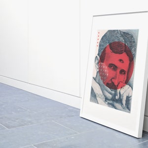 May include: A framed print of a portrait of Nikola Tesla with a red circle over his face. The circle has a third eye in the centre. The print is in a white frame and is leaning against a white wall.