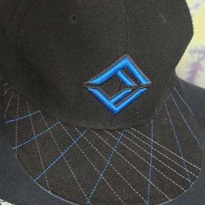 Puede incluir: Gorra de béisbol negra con un logotipo bordado en azul y blanco. La visera presenta un patrón geométrico con costuras azules. La gorra está hecha de una tela texturizada y tiene una corona redondeada.