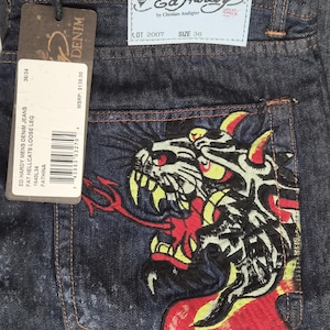 Puede incluir: Vaqueros de mezclilla azul oscuro con un gran gráfico bordado y colorido de un gato estilizado en el bolsillo trasero. Los vaqueros incluyen una etiqueta de marca azul claro con el texto Ed Hardy by Christian Audigier y una etiqueta colgante.