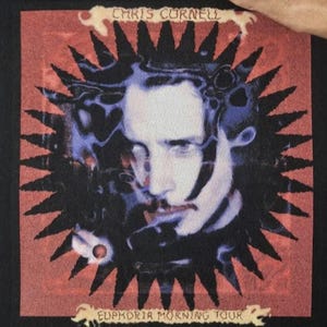 May include: A black t-shirt featuring a graphic print of Chris Cornell. The design includes a stylized portrait surrounded by a black sunburst shape on a red background. The text "CHRIS CORNELL" appears at the top and "EUPHORIA MORNING TOUR" at the bottom.