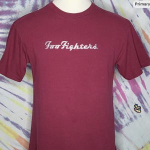 Puede incluir: Una camiseta burdeos de manga corta con el texto "Foo Fighters" en plata y cursiva en el pecho. La camiseta se muestra en un maniquí sobre un fondo tie-dye colorido.