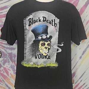 Puede incluir: Camiseta negra con un gráfico de una calavera con un sombrero de copa y fumando un cigarrillo. La calavera está delante de una lápida que dice "Black Death Vodka".