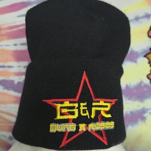 Puede incluir: Gorro de punto negro con una estrella roja y texto amarillo y gris. El texto dice "Guns N Roses". Un accesorio de invierno clásico.
