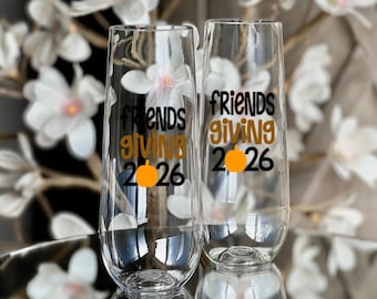 May include: Two clear stemless champagne flutes featuring black and orange text that reads friends giving 2026, with an orange pumpkin icon replacing the letter i in the word giving and the number zero in the year 2026.