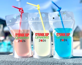 May include: Three clear plastic drink pouches with straws, each filled with a different colored liquid: pink, yellow, and blue. The pouches feature the red and green text "DRINK UP Grinches 2026" with a red ornament replacing the zero in 2026.