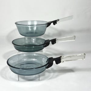 Può includere: Tre padelle vintage in vetro grigio chiaro con manici neri e impugnature in plastica trasparente. Le padelle sono impilate, mostrando la loro forma rotonda e il design dei manici. Il vetro ha una leggera sfumatura blu-grigia.