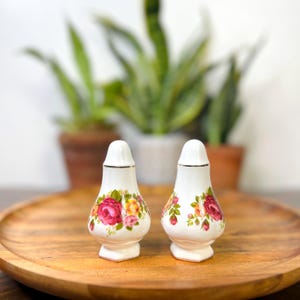 Puede incluir: Un par de saleros y pimenteros de cerámica blanca con un diseño floral de rosas. Los saleros y pimenteros tienen un borde dorado y se exhiben en una bandeja de madera. Las rosas son rosas, amarillas y rojas.