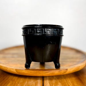 Puede incluir: Un recipiente decorativo negro de tres patas con un diseño de clave griega alrededor del borde. El recipiente está sobre una bandeja redonda de madera. El recipiente tiene un acabado brillante y un diseño simple y elegante.