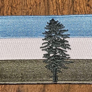 May include: An embroidered patch featuring a blue, white and green flag with a black silhouette of a pine tree in the centre.
