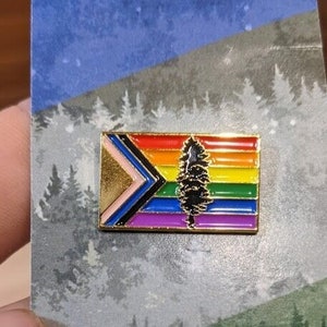 May include: A gold-colored pin with a rainbow flag design featuring a stylized evergreen tree in the center. The flag has a pink, blue, white, black, and purple stripe pattern.