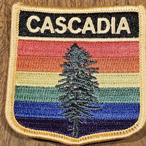 May include: A colorful embroidered patch with a rainbow design and a stylized evergreen tree. The patch has the word "Cascadia" in white text on a black background.