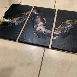 May include: A three-piece abstract art canvas set with black, gold, white, and orange paint. The abstract design is reminiscent of a flowing river or stream.