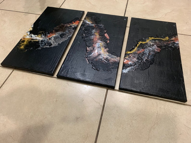 May include: A three-piece abstract art canvas set with black, gold, white, and orange paint. The abstract design is reminiscent of a flowing river or stream.