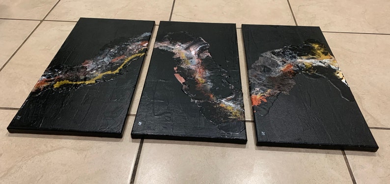 May include: Three black canvas paintings with abstract designs in gold, white, and orange. The designs are reminiscent of flowing water or a river.
