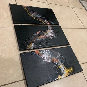 May include: Three black canvas panels with abstract art featuring gold, white, and pink paint. The panels are arranged horizontally and are ready to be hung on a wall.