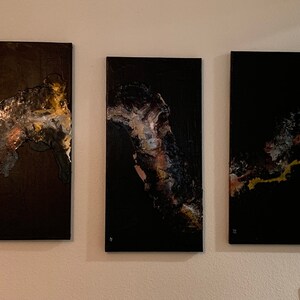 May include: Three abstract paintings on canvas with black backgrounds and gold, silver, and brown accents. The paintings are arranged in a row on a white wall.