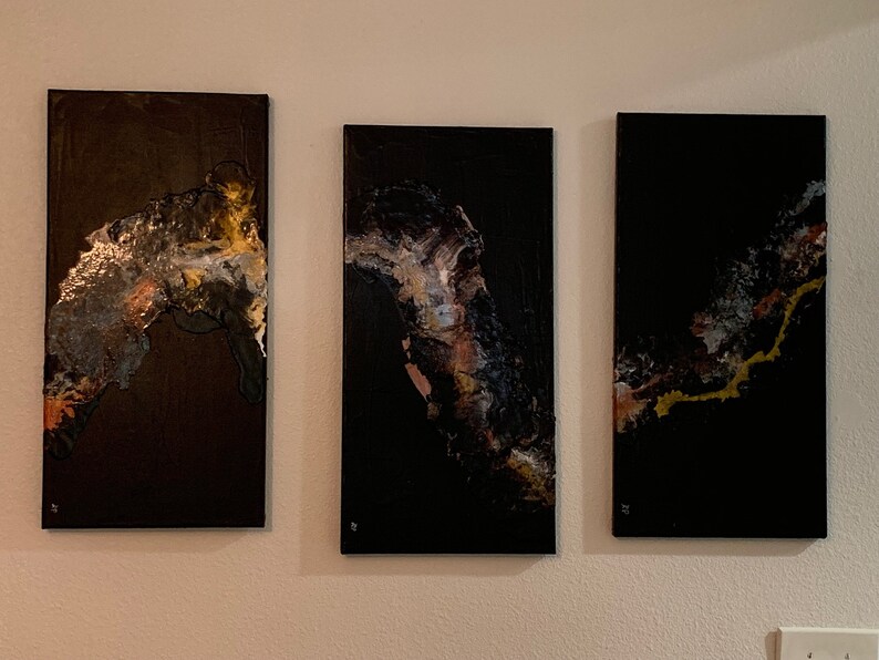 May include: Three abstract paintings on canvas with black backgrounds and gold, silver, and brown accents. The paintings are arranged in a row on a white wall.