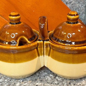 Puede incluir: Un servidor de condimentos de cerámica de dos secciones con asa, en marrón y crema. Cada sección tiene una tapa con un pomo. Una sección tiene una cuchara. El servidor es probablemente para sal, pimienta u otras especias.