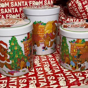 May include: Three white metal Christmas tins with bear illustrations. Each tin features a different scene with bears, Christmas trees, and fireplaces. The tins are on a red and white patterned wrapping paper with the words "Santa From Santa" printed on it.