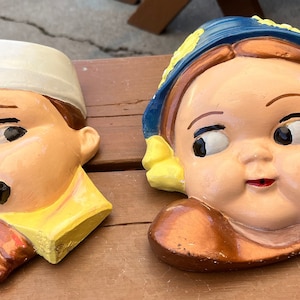 May include: Two vintage ceramic wall hangings depicting cartoon-style faces. One features a boy in a white hat and yellow collar, the other a girl in a blue hat with yellow accents. Both have rosy cheeks and painted details.