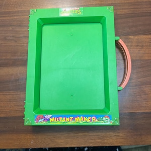 May include: A green plastic toy with a rectangular frame and a handle. The toy features the words "Mutant Maker" and "Turtles". The handle is a reddish-brown colour.