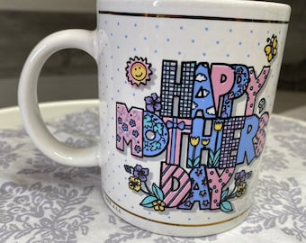 Könnte beinhalten: Weiße Keramik-Tasse mit der Aufschrift "HAPPY MOTHER'S DAY" in bunten, gemusterten Buchstaben. Die Tasse hat einen Henkel, einen goldenen Rand und ist mit kleinen blauen Punkten, Blumen, einer Sonne und einem Schmetterling verziert.