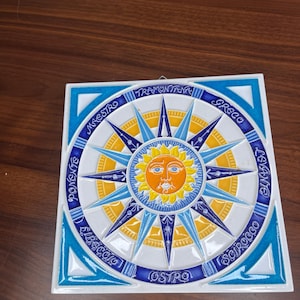 May include: Square ceramic tile featuring a compass rose design with a sun in the center. The tile has a white background with blue and yellow accents. The compass points are labeled with Italian words. The tile is approximately 8 inches square.