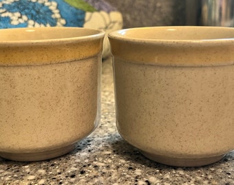 May include: Two beige ceramic mugs with a speckled design and a yellow rim. One mug has a handle. The mugs are sitting on a speckled countertop. The mugs are likely for coffee or tea.