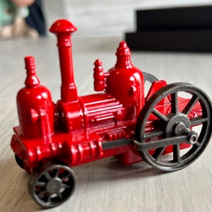 May include: A small, red toy steam engine with black wheels and a tall smokestack. The miniature engine has detailed features, including a boiler, cylinders, and a flywheel. The toy is a vintage-style collectible.