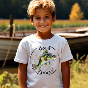 May include: White t-shirt featuring a fish graphic and the text "Gone Fishin'". A child in blue jeans smiles in front of a lake with a boat. The image captures a sunny day and the theme of fishing.