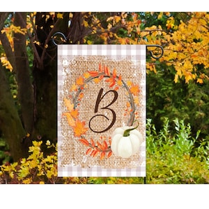 May include: A garden flag featuring a burlap texture background with a wreath of fall leaves and a white pumpkin. The letter "B" is centered within the wreath. The flag has a tan and white checkered border.